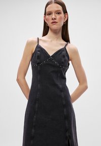 Vestido de ganga preto com alças finas, decote em V e detalhes decorativos de tachas na zona do peito. Silhueta ajustada com racha frontal.