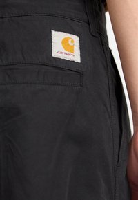 Czarne ogrodniczki z białą kwadratową łatą, na której znajduje się pomarańczowe logo i czerwony napis. Materiał wygląda na gładki, z widocznymi szwami i kieszeniami.