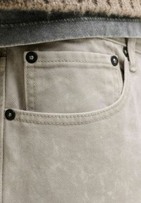 Μπεζ τζιν παντελόνι με μαύρα καρφιά και μια μικρή τσέπη, φορεμένο με γκρι πουκάμισο και μπεζ πλεκτό πουλόβερ.