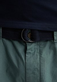 Ceinture tissée bleu marine avec boucle double D en métal, portée avec un pantalon vert et une chemise bleu foncé rentrée.