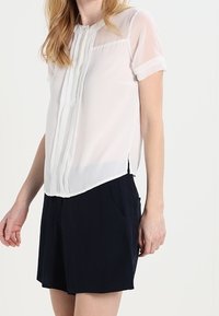 Blusa bianca a maniche corte con fronte plissettato, tessuto trasparente e orlo arrotondato. Abbinata a pantaloni corti blu scuro con tasche laterali.