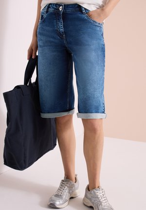 Short en denim bleu longueur genou avec revers roulés porté avec des baskets argentées ; personne tenant un grand sac cabas noir, main gauche dans la poche.