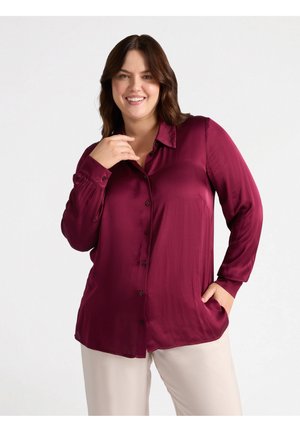 Camicia button-up in satin bordeaux con colletto, maniche lunghe e vestibilità rilassata. Presenta una tasca frontale e una texture liscia. Indossata con pantaloni chiari.