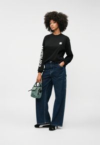 Melns ilgais krekls ar logo, kombinēts ar brīviem zila džinsiem. Tur rokās maza zaļa soma, uz kājām melni slip-on apavi. Vienkārša fona.