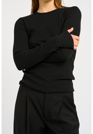 Femme portant un haut côtelé noir à manches longues avec un petit logo sur la poitrine, les bras croisés, assorti à un pantalon noir.