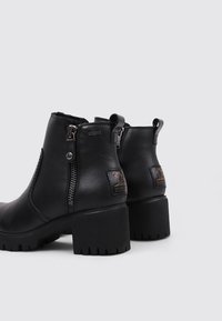 Botas de tobillo de cuero negro con un acabado texturizado, que cuentan con un cierre lateral y un tacón grueso. Acentuadas con una etiqueta de la marca en la parte trasera.