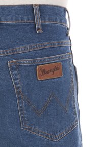 Jeans in denim blu con un'etichetta in pelle marrone che presenta la scritta "Wrangler". Design notevole della tasca posteriore con un motivo a zigzag.