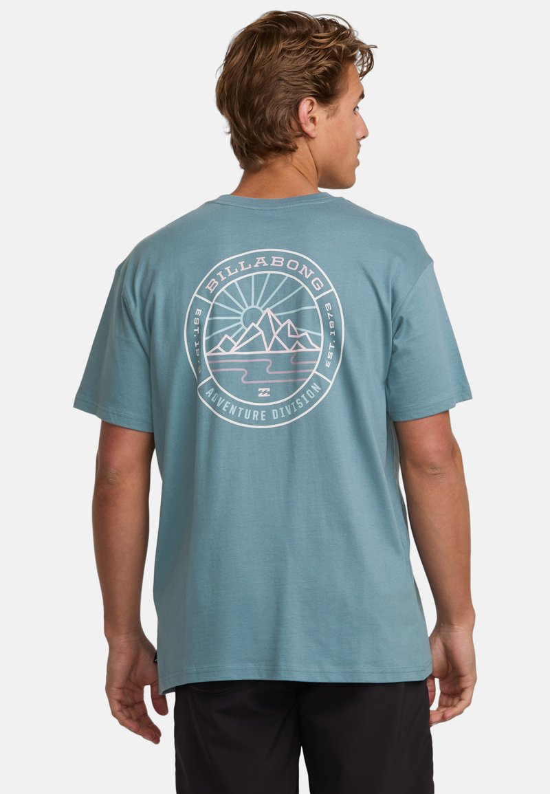 Light teal cotton t-shirt featuring a circular graphic with mountains and sun illustration in white and pink, labeled "Billabong Adventure Division."