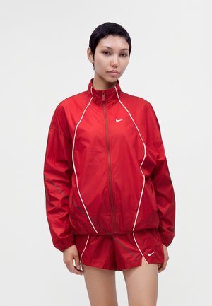 Femme portant une veste zippée Nike rouge et un short rouge assorti avec un bord blanc, debout devant un fond clair uni.