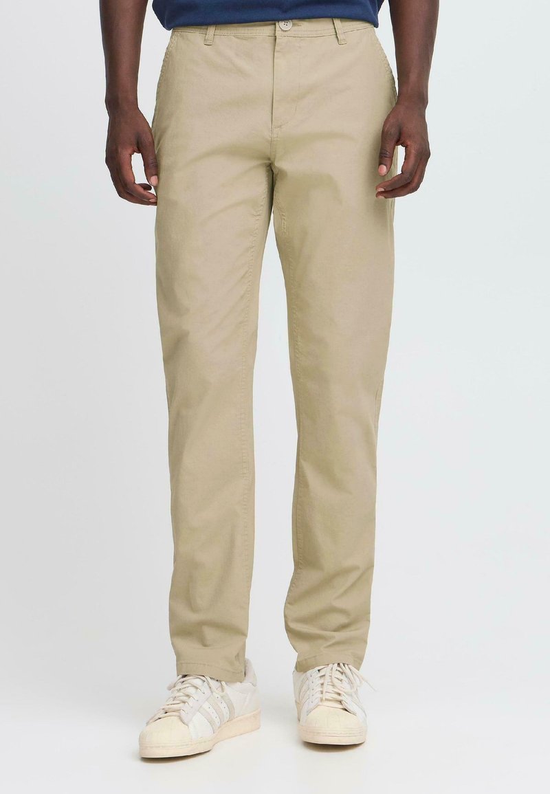Tan cotton chinos with a straight-leg design, featuring a front button closure and side pockets, paired with off-white sneakers.