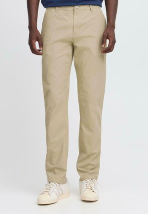 Man som bär beige raka byxor och vita sneakers som står mot en enkel ljus bakgrund.