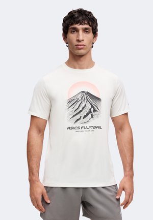 Jeune homme debout, portant un t-shirt blanc avec un motif de montagne noir et rouge et un short gris sur un fond clair uni.