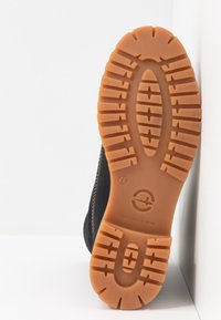 Chaussures noires avec une semelle en caoutchouc présentant des crampons texturés et un bord cousu, affichant une semelle intermédiaire marron et un logo circulaire.