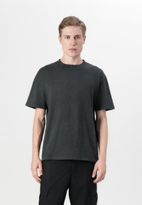 Νεαρός άνδρας που στέκεται με το πρόσωπο προς τα εμπρός, φορώντας ένα απλό σκουρό γκρι κοντομάνικο μπλουζάκι και μαύρο παντελόνι φόρμας, μπροστά σ ε ένα φωτεινό φόντο.