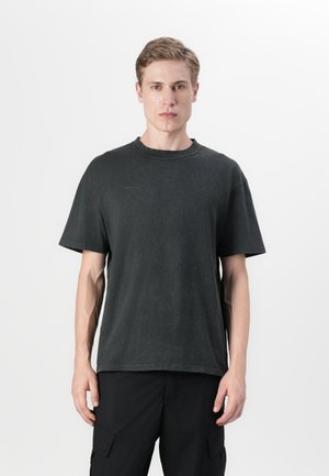 Ung mand står med front mod kameraet iført en ensfarvet mørkegrå kortærmet t-shirt og sorte cargobukser foran en lys baggrund.