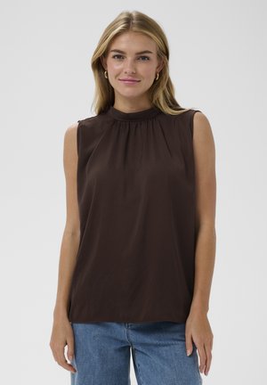 Giovane donna con capelli biondi che indossa orecchini a cerchio dorati, top marrone senza maniche con scollo arricciato e jeans blu su sfondo semplice.