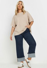 Camiseta beis de gran tamaño con mangas cortas, combinada con vaqueros de pierna ancha de color azul oscuro con un dobladillo de denim. Zapatillas blancas con rayas negras.