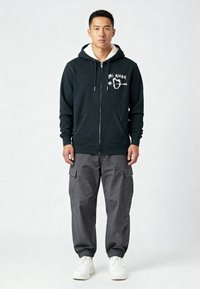 Zwarte zip-up hoodie met een fleece gevoerde kraag, voorzien van witte tekstgraphics. Gepaarde met grijze cargobroek en witte sneakers.