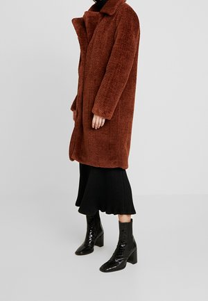 Femme portant un long manteau marron duveteux, une jupe midi noire côtelée et des bottines noires brillantes, debout sur un fond clair uni.