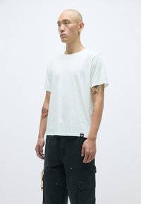 Bijela pamučna majica klasičnog kroja s okruglim izrezom i kratkim rukavima, u kombinaciji s crnim cargo hlačama koje imaju džepove i suptilne metalne detalje.