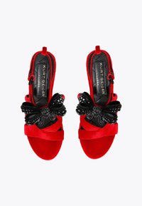 Czerwone zamszowe sandały na wysokim obcasie z odkrytymi palcami, ozdobione czarnymi kokardkami i kryształowymi detalami, z krzyżującymi się paskami i konstrukcją z zapięciem na pięcie.