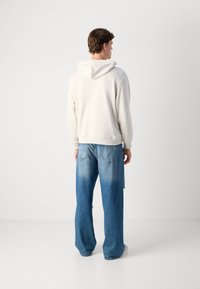 Helle beige Kapuzenpulli mit Kordelzug, kombiniert mit weit geschnittenen blauen Jeans mit einem verwaschenen Design und Gesäßtaschen, vor einem schlichten Hintergrund.