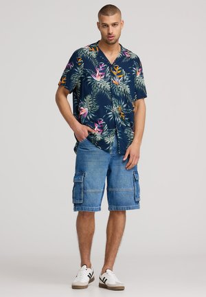 Camicia a maniche corte blu navy con stampa floreale tropicale, abbinata a pantaloni cargo in denim e sneakers bianche con strisce nere. Vestibilità casual.