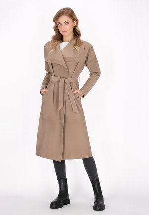 Vrouw draagt een lange beige mantel met brede revers en ceintuur, zwarte legging en zwarte grove laarzen, staat met handen in de zakken.