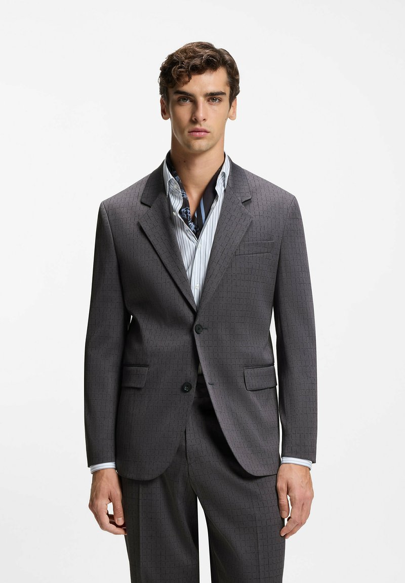 Veste de costume grise texturée avec deux poches avant, un bouton unique et un revers cranté. Portée sur une chemise rayée et une écharpe à motifs.