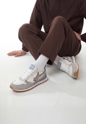 Persona con pantalones marrones, calcetines blancos y zapatillas blancas con detalles en beige y gris, sentada en el suelo con las piernas cruzadas.