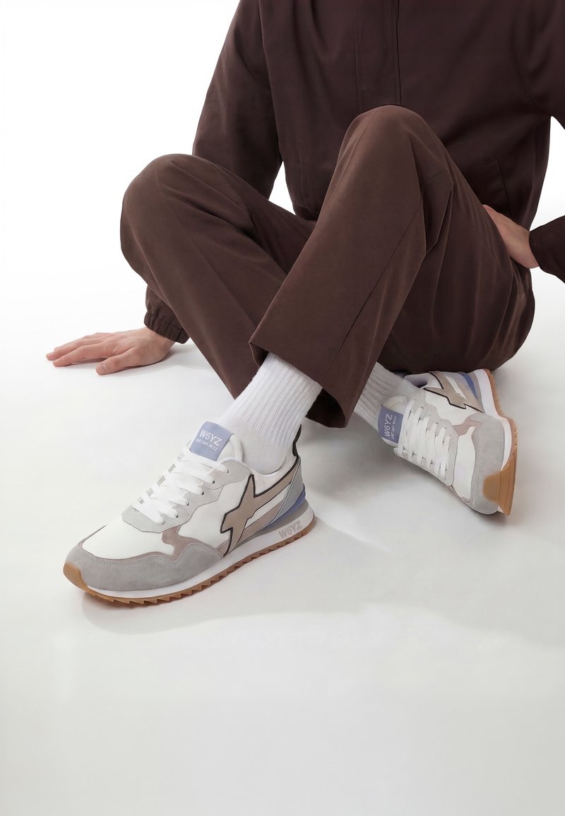 Personne portant un pantalon marron, des chaussettes blanches et des baskets blanches avec des touches beige et grises, assise par terre, jambes croisées.