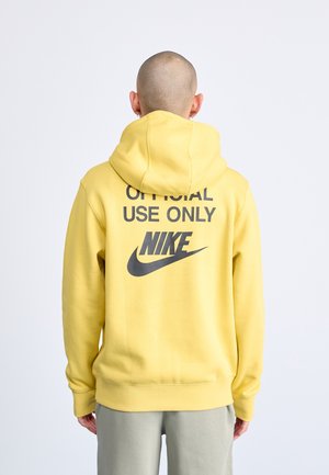 Person med barberet hoved iført gul Nike-hættetrøje med teksten "Official Use Only" på ryggen, stående mod en ensartet lys baggrund.