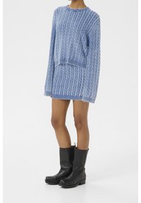Pull en tricot bleu clair avec motif torsadé, associé à une jupe assortie bleu clair. Bottes hautes en cuir noir avec semelles épaisses.