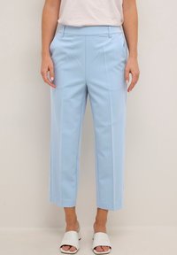 Donna che indossa pantaloni su misura azzurri tagliati con cuciture frontali, abbinati a sandali bianchi con tacco aperto e a un top bianco.