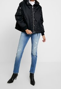 Giacca a vento nera con cappuccio e chiusura a zip, con tasche laterali e polsini a coste. Abbinata a jeans skinny blu strappati e tacchi neri.