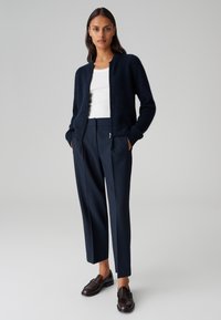 Giacca bomber lavorata a maglia blu navy con polsini a costine, indossata sopra un top bianco, abbinata a un paio di pantaloni navy a vita alta e mocassini in pelle scura.