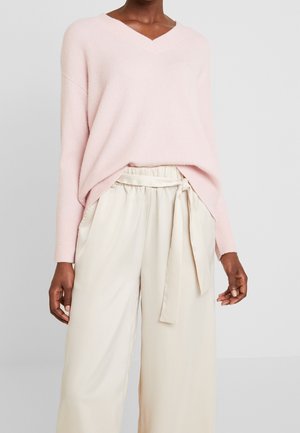 Femme portant un pull à col en V rose clair et un pantalon paperbag crème taille haute avec une ceinture nouée.