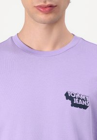 Lavandos spalvos medvilninis T-shirt su apvaliu kaklu ir trumpomis rankovėmis. Kairėje pusėje yra juodas "TOMMY JEANS" logotipas. Slydus paviršius.