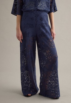 Pantaloni a gamba larga blu navy con motivi traforati in pizzo floreale, indossati con top abbinato e sandali neri aperti sulle dita.