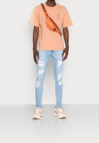 Peach short-sleeve t-shirt with a chest graphic, light blue ripped skinny jeans, orange sling bag, and beige athletic shoes with orange accents.