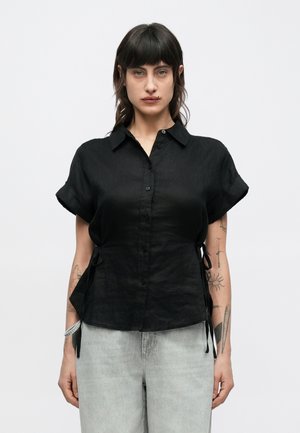 Fekete lenből készült rövid ujjú ing gombos elülső résszel, lekerekített aljjal és oldalsó megkötőkkel az állítható illeszkedésért. Sima textúra, enyhe fényű felülettel.