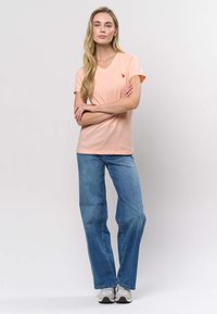 Hellpfirsichfarbenes V-Ausschnitt-T-Shirt mit kurzen Ärmeln, kombiniert mit locker sitzenden blauen Jeans. Das T-Shirt hat ein kleines rotes Logo auf der Brust.