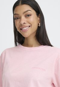 Lächelnde Frau mit langen dunklen Haaren, die silberne Creolen und ein hellrosa Hemd trägt, auf dem "IMPERFECTION" auf der Brust gedruckt ist.