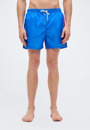 Mann trägt leuchtend blaue Badeshorts mit weißem Kordelzug und rotem Seitenlogo, steht barfuß vor einfarbigem Hintergrund.