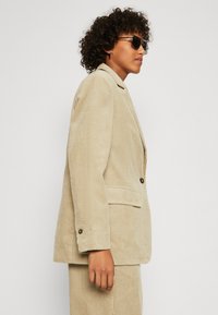 Beige kordfløjl blazer med notched krave, to forlommer og knaplukning. Mønstret stof med lodrette striber og skræddersyet pasform.