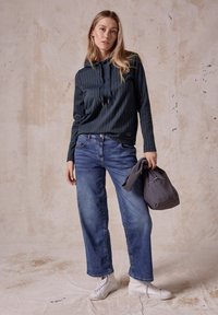 Navy gestreifter Kapuzenpullover mit verstellbaren Kordeln, kombiniert mit lockeren blauen Jeans und weißen High-Top-Sneakern. Graue Handtasche in der Hand.