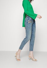 Camisa de manga larga texturizada verde, Jeans de tiro alto azul claro con bolsillos con cremallera y sandalias de tacón con tiras beige sobre un fondo liso.
