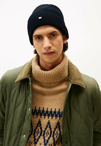 Beanie lavorato a maglia blu scuro, maglione a collo alto a coste color sabbia con motivo geometrico blu, giacca verde oliva trapuntata con bottoni a pressione.