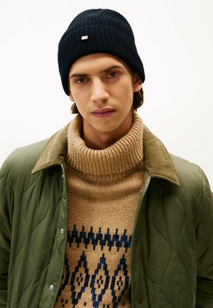 Beanie lavorato a maglia blu scuro, maglione a collo alto a coste color sabbia con motivo geometrico blu, giacca verde oliva trapuntata con bottoni a pressione.