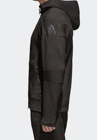 Svart zip-up hoodie i strukturerad tyg, med huva, sidofickor med dragkedja och axellogo i matt finish.